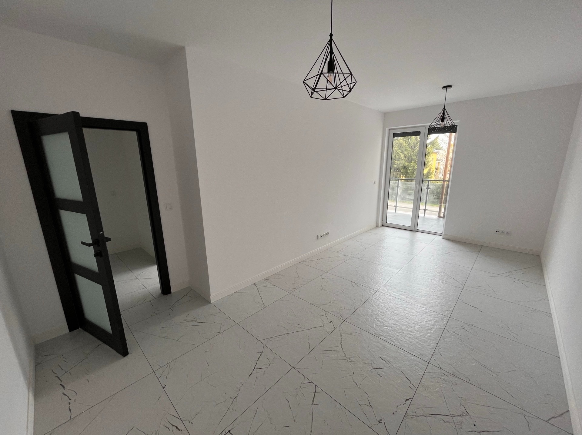 Jasne wnętrze z marmurową podłogą i otwartymi, czarnymi drzwiami. Geometryczne lampy wiszące dodają nowoczesnego charakteru. Widok na balkon przez przeszklone drzwi.