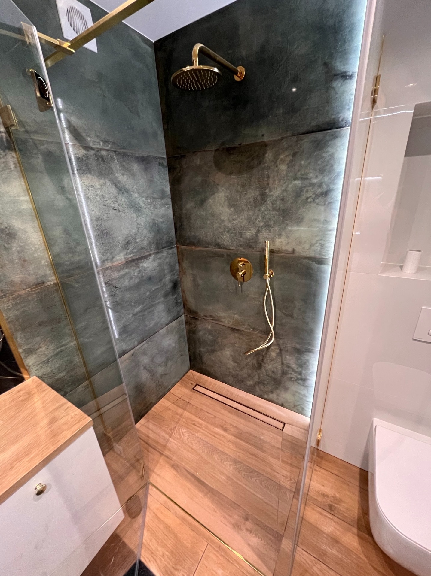 Nowoczesna kabina prysznicowa z zielonymi płytkami imitującymi kamień, złotą armaturą i drewnianą podłogą. Minimalistyczny design z subtelnym oświetleniem LED.
