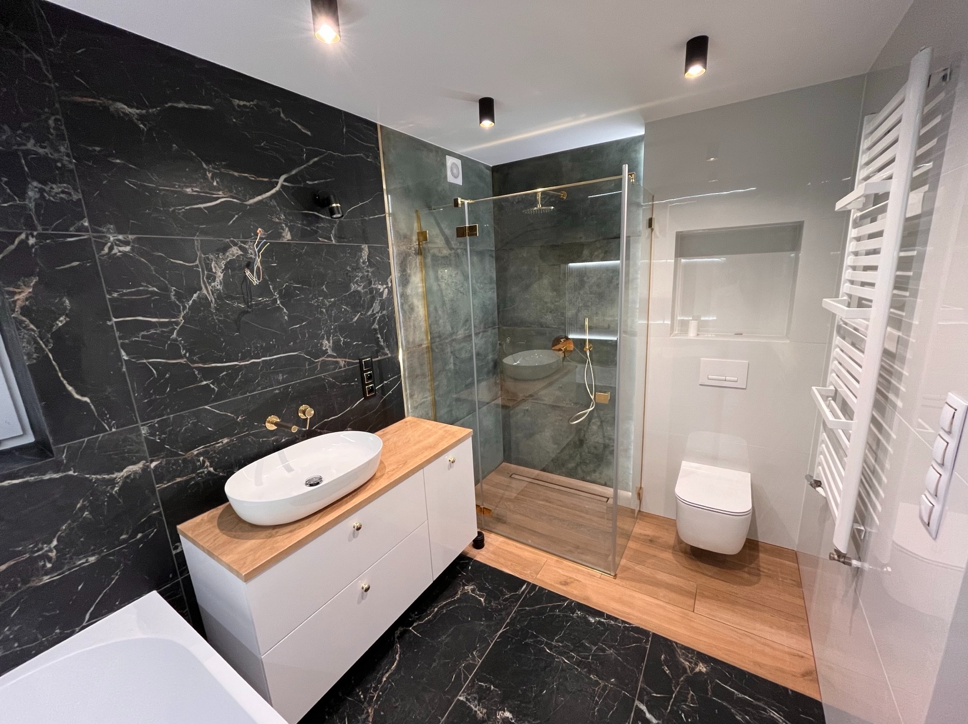 Nowoczesna łazienka z czarnymi marmurowymi płytkami, kabiną prysznicową typu walk-in i białą umywalką nablatową na szafce z drewnianym blatem.
