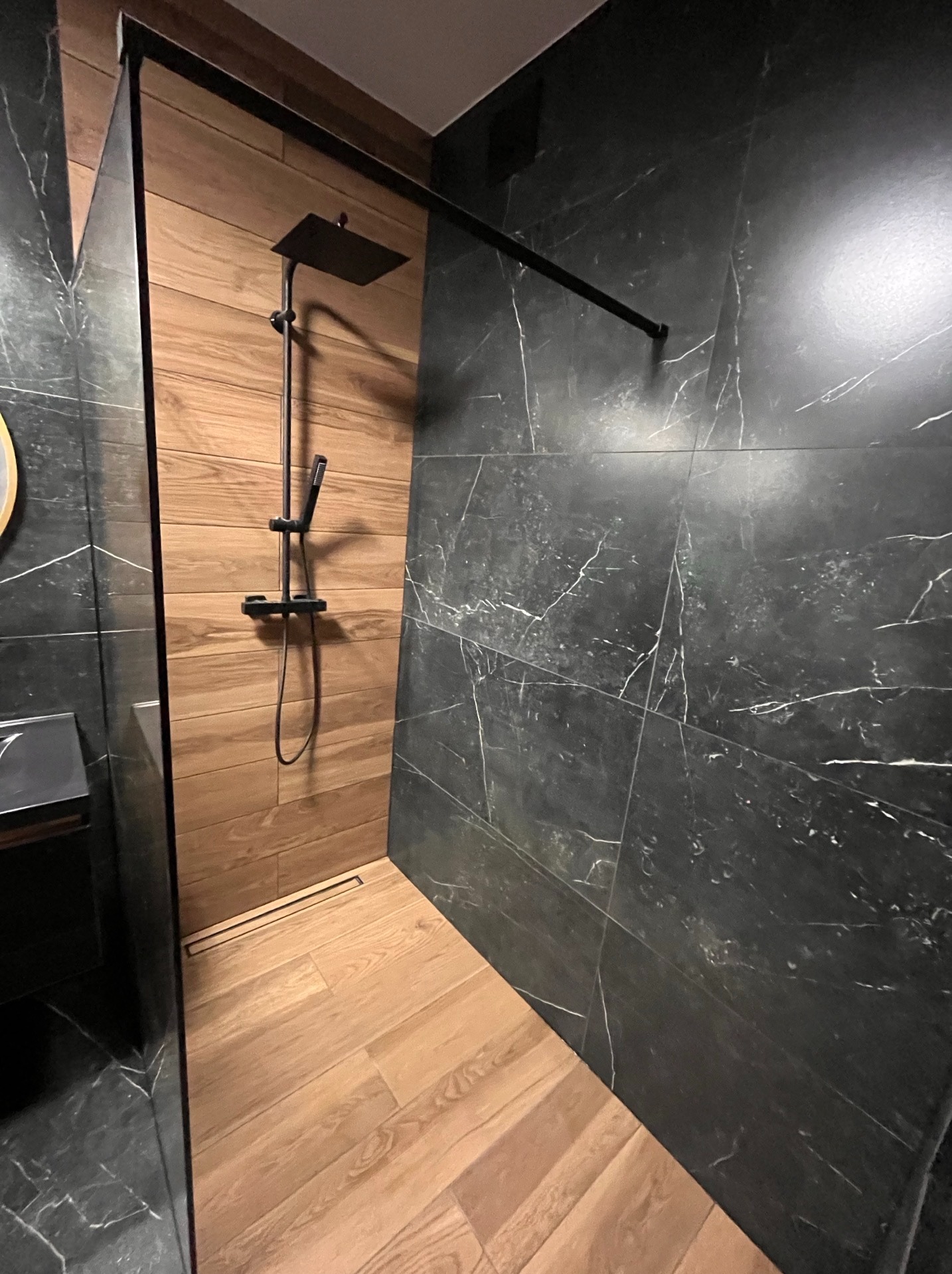 Nowoczesna kabina prysznicowa z czarną armaturą i płytkami imitującymi drewno oraz marmur, z odpływem liniowym w podłodze. Minimalistyczny design.