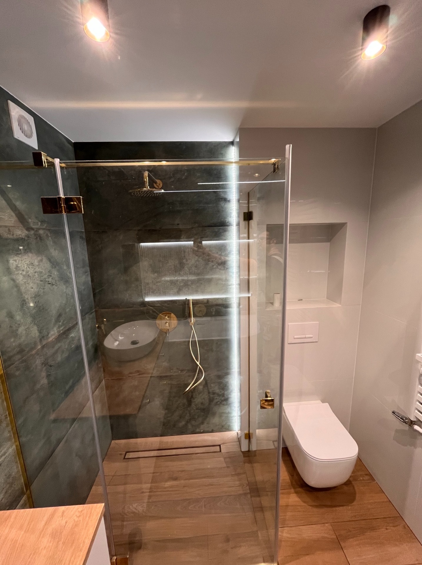 Nowoczesna łazienka z kabiną prysznicową typu walk-in, wykończona płytkami imitującymi kamień i drewnem. Złote detale armatury i oświetlenia dodają elegancji.