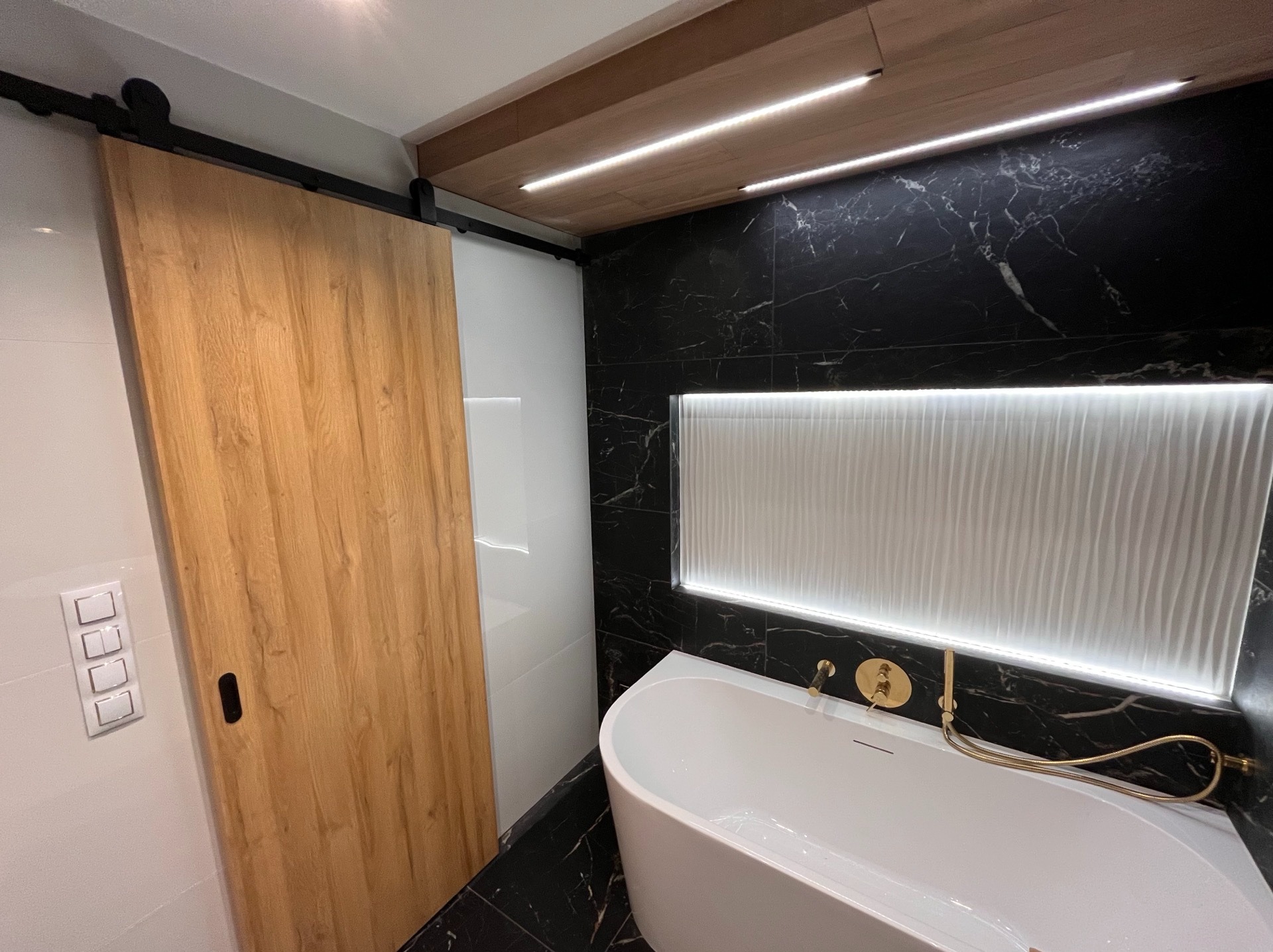Nowoczesna łazienka z przesuwnymi drzwiami z jasnego drewna, czarnymi marmurowymi płytkami i podświetlaną wanną. Minimalistyczny design z akcentami złota.