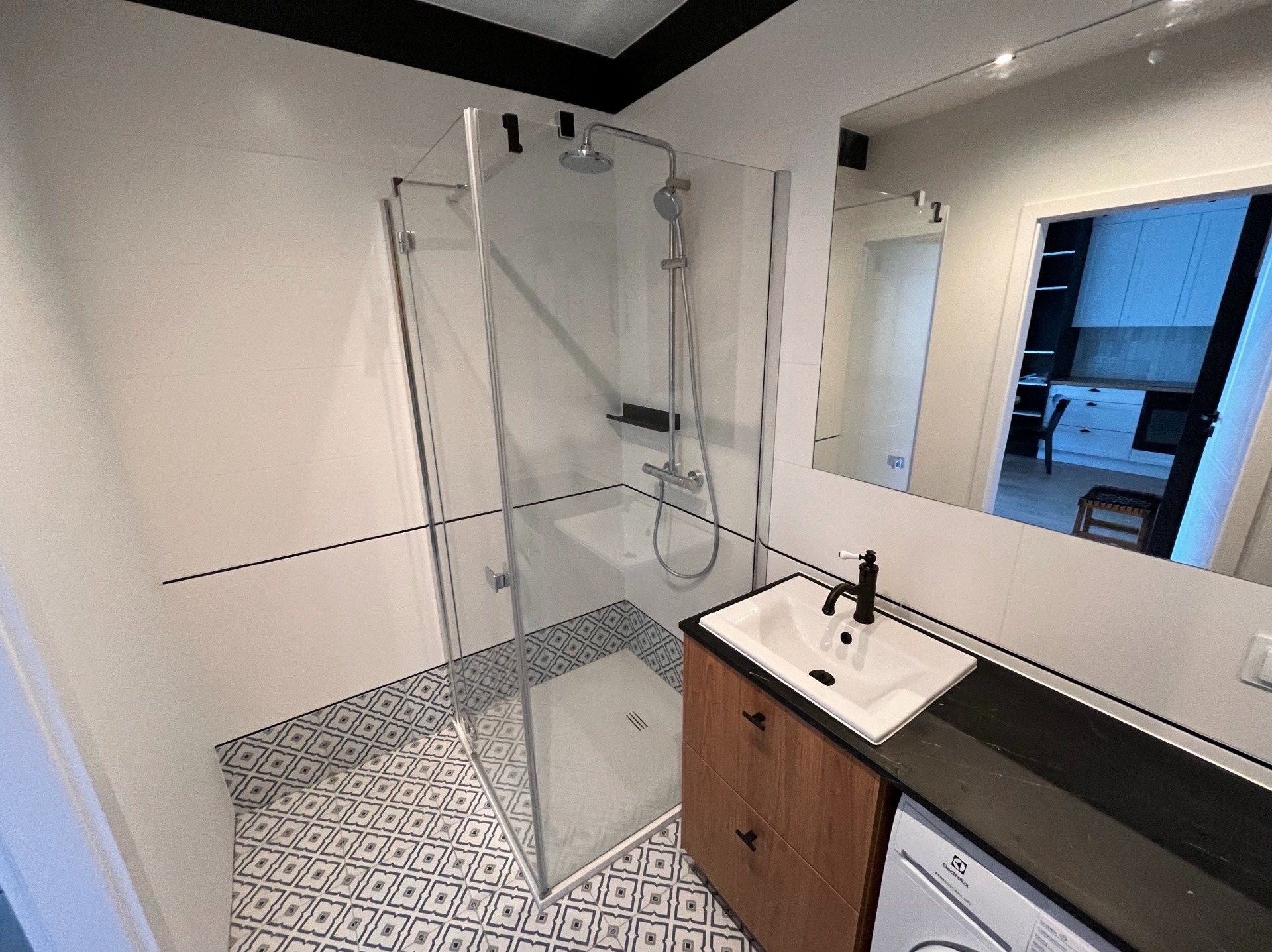 Nowoczesna łazienka z kabiną prysznicową typu walk-in, białą umywalką na szafce z drewnianymi frontami i wzorzystą podłogą. W tle widoczne lustro i fragment kuchni.