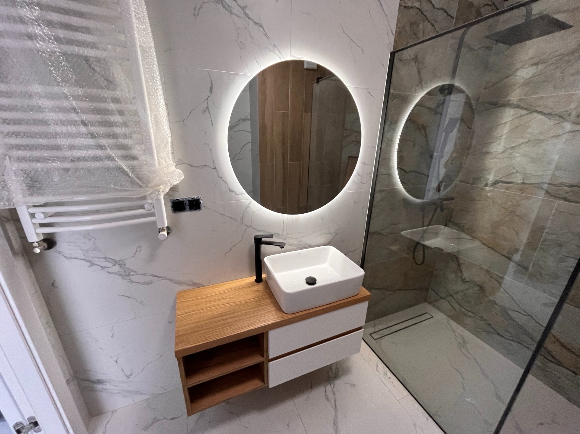 Nowoczesna łazienka z marmurowymi płytkami, okrągłym lustrem z podświetleniem LED, drewnianą szafką pod umywalką i kabiną prysznicową ze szklaną ścianą.