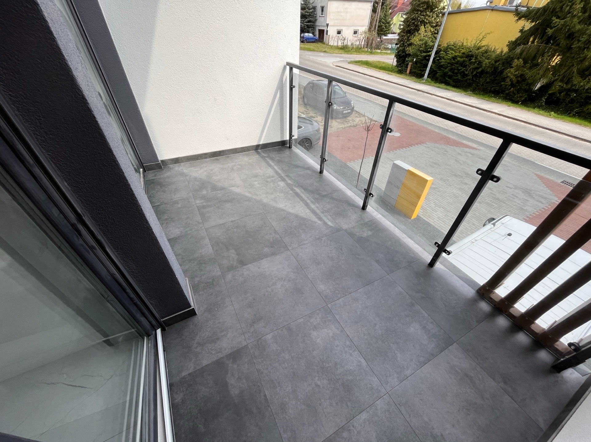 Wykończony balkon z szarymi płytkami i szklaną balustradą w Łężycy. Widok na ulicę. Nowoczesny design, staranne ułożenie płytek, czysty i estetyczny wygląd.