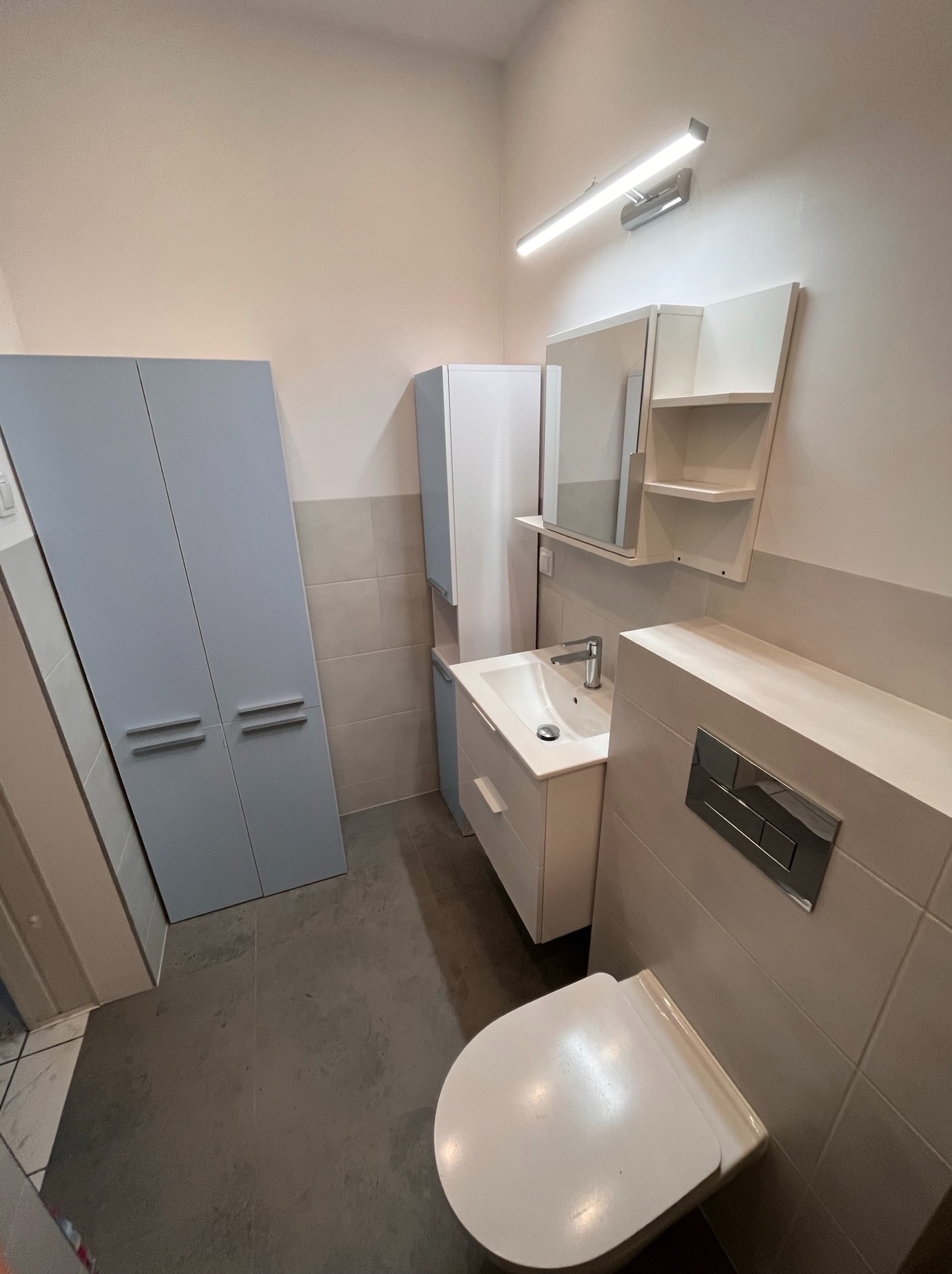 Nowoczesna łazienka po remoncie: szara podłoga, białe meble łazienkowe, podwieszana toaleta z szarymi płytkami. Minimalistyczny design i oświetlenie.