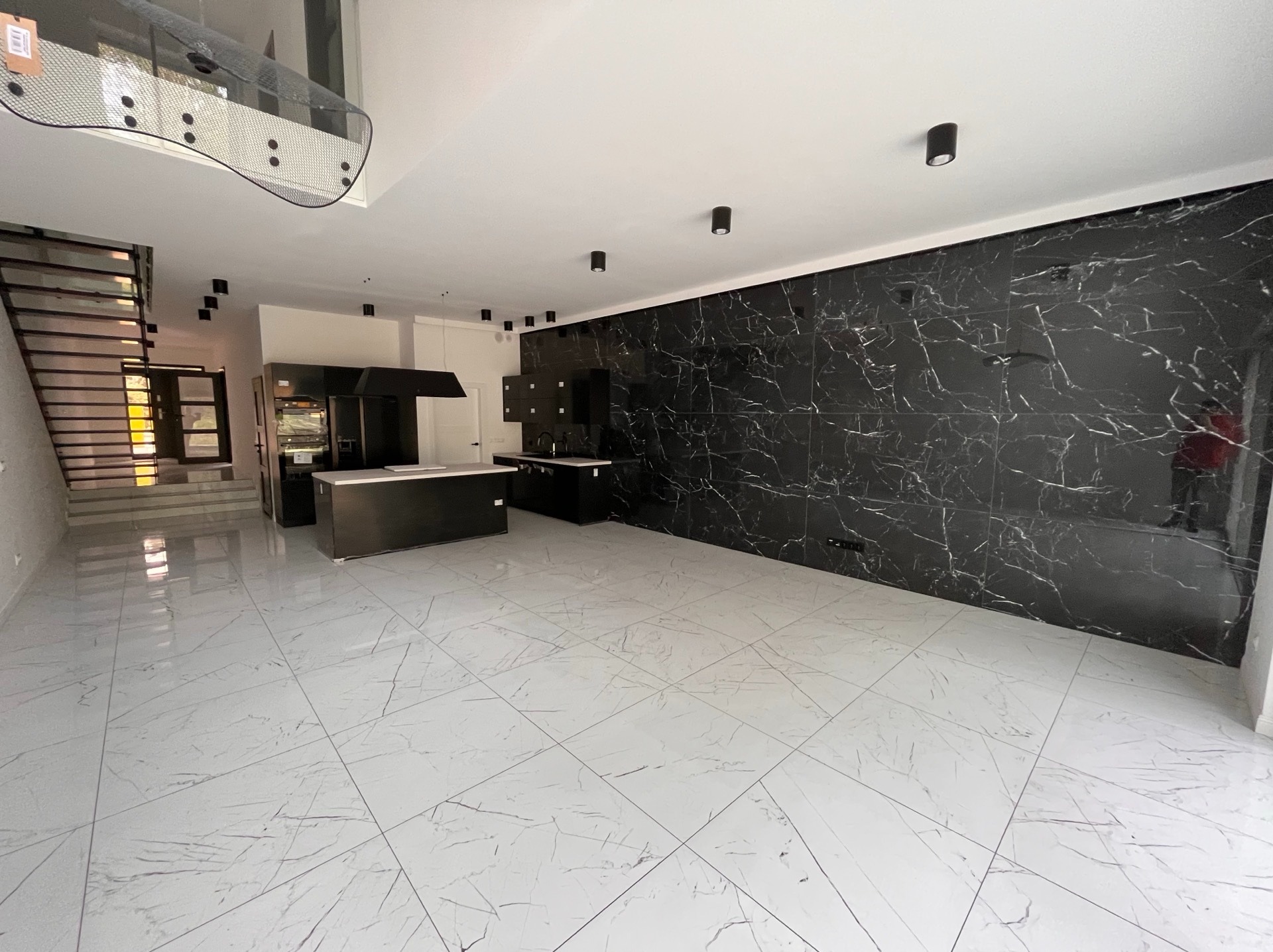 Nowoczesne wnętrze z marmurową ścianą i podłogą. Minimalistyczna kuchnia z czarnymi szafkami i wyspą. Schody z metalową balustradą. Oświetlenie punktowe.