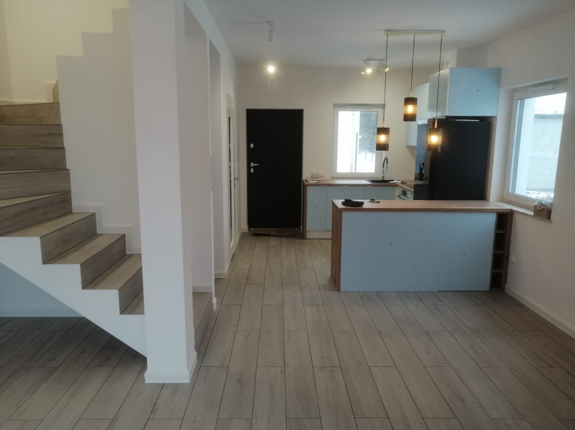 Jasne wnętrze domu po remoncie: schody, panele podłogowe, kuchnia z wyspą i oświetleniem. Minimalistyczny design, neutralna kolorystyka, widok na drzwi wejściowe.