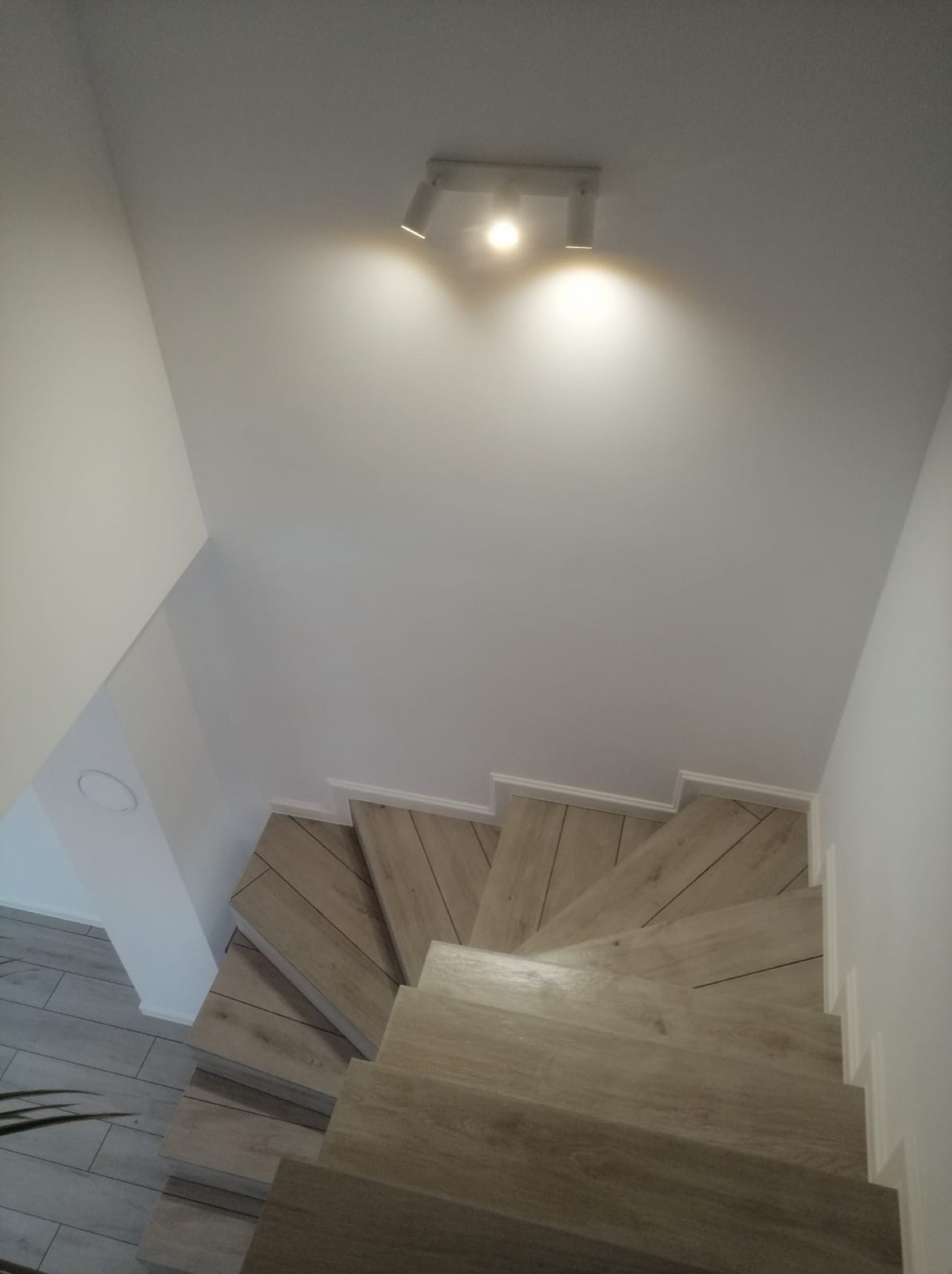 Wykończone schody z drewnopodobnych paneli, oświetlone punktowym światłem. Białe ściany i listwy przypodłogowe. Widok z góry, podkreślający estetykę wykończenia.