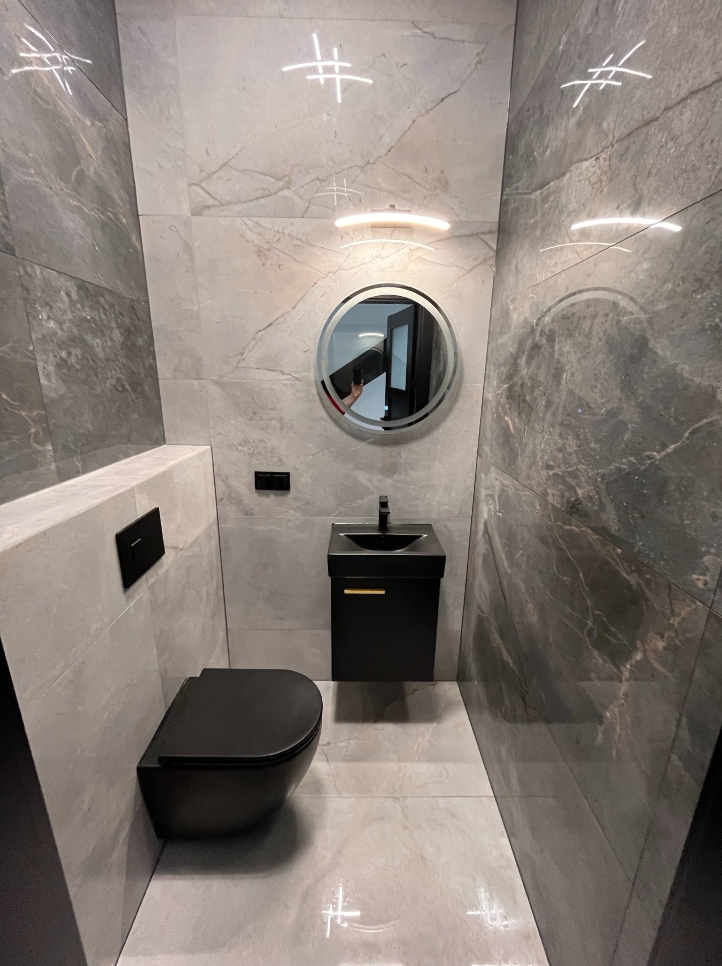 Nowoczesna, mała łazienka w szarościach z podwieszaną toaletą i okrągłym lustrem z podświetleniem. Minimalistyczny design, marmurowe płytki, czarna armatura.