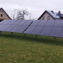 EMPA Sp z o.o - Dwa rzędy paneli słonecznych na trawniku przed domami jednorodzinnymi, część paneli zamontowana również na dachu jednego z budynków.