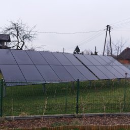 EMPA Sp z o.o - Rząd paneli słonecznych na metalowej konstrukcji, widoczny zza niskiego, zielonego płotu na tle domu i pochmurnego nieba.