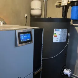 Szary piec z dotykowym wyświetlaczem wskazującym 45 stopni Celsjusza, obok czarny zbiornik z etykietą energetyczną w kotłowni.