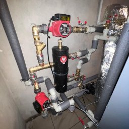 EMPA Sp z o.o - Złożona instalacja hydrauliczna z dwoma czerwonymi pompami obiegowymi marki Ferro, czarnym zbiornikiem z naklejką z orłem w koronie, rurami miedzianymi i izolowanymi rurami PCV, zamontowana...