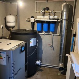 EMPA Sp z o.o - Kotłownia z kotłem na pellet, zbiornikiem buforowym i filtrami wody, widoczne rury instalacji grzewczej, szara ściana i brązowa podłoga z płytek.