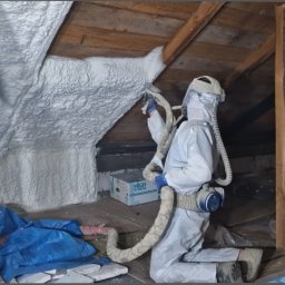 EMPA Sp z o.o - Ocieplanie poddasza pianką poliuretanową przez pracownika w kombinezonie ochronnym i masce z filtrem, widoczna struktura pianki i drewniane elementy konstrukcyjne dachu.