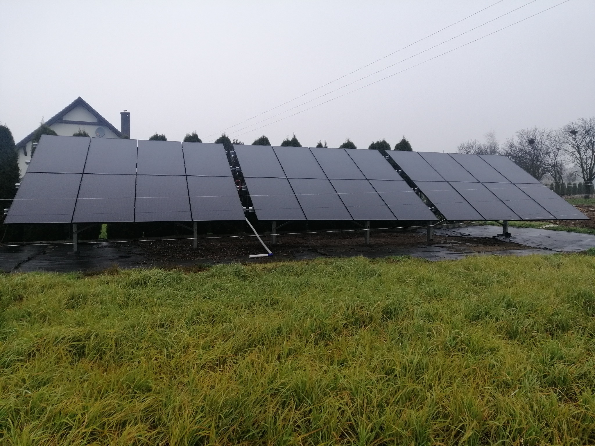 Instalacja fotowoltaiczna na trawiastym terenie w pochmurny dzień, widoczny dom w tle.