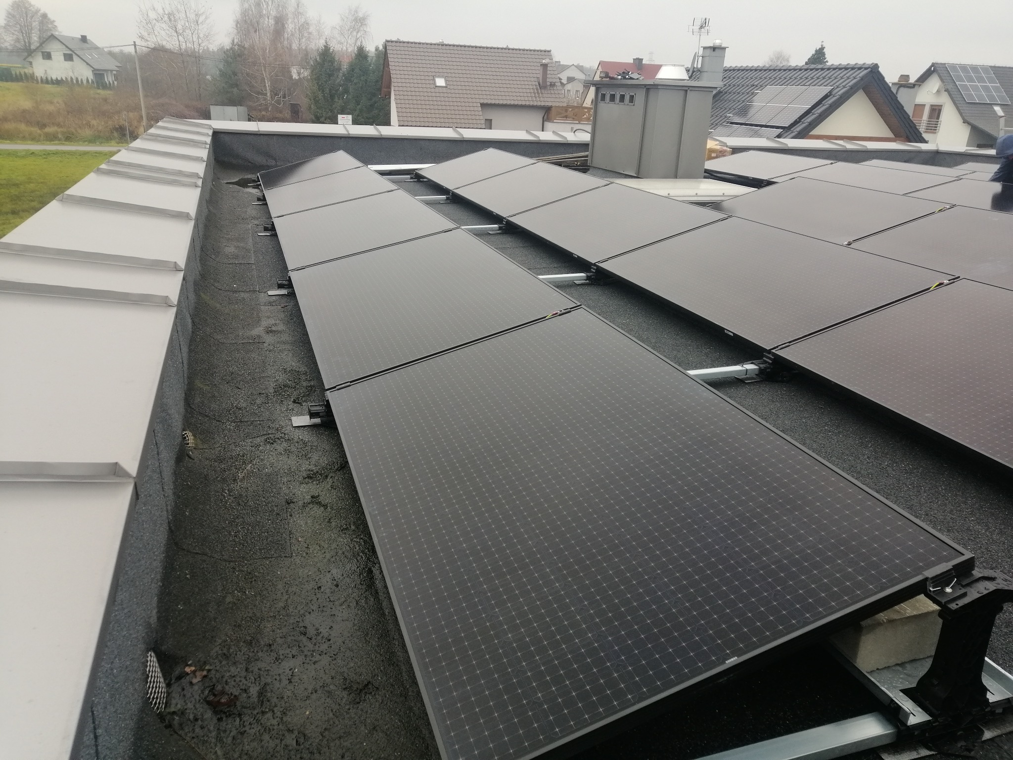 Rząd paneli słonecznych na płaskim dachu z widocznym systemem montażowym, w tle zabudowa jednorodzinna w pochmurny dzień.