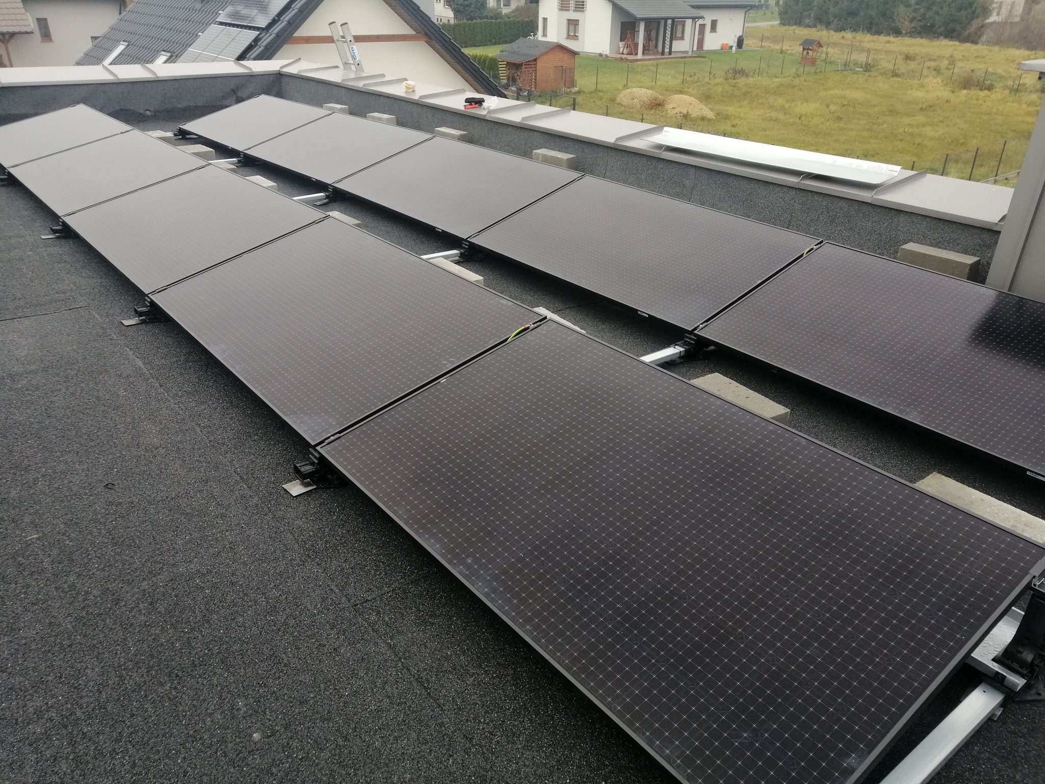 Rząd paneli słonecznych zamontowanych na płaskim dachu pokrytym papą, widoczne elementy montażowe i fragment otoczenia z domami.