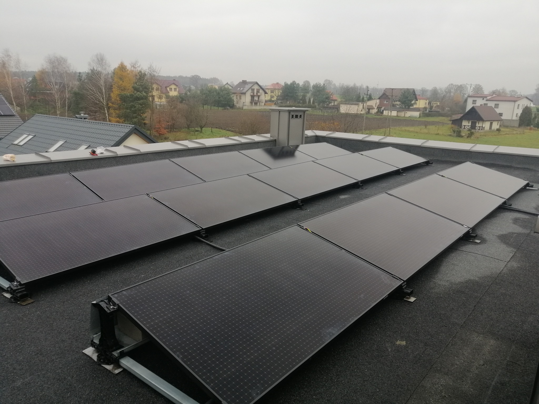 Rząd paneli fotowoltaicznych na płaskim dachu z widokiem na wiejskie domy w pochmurny dzień.