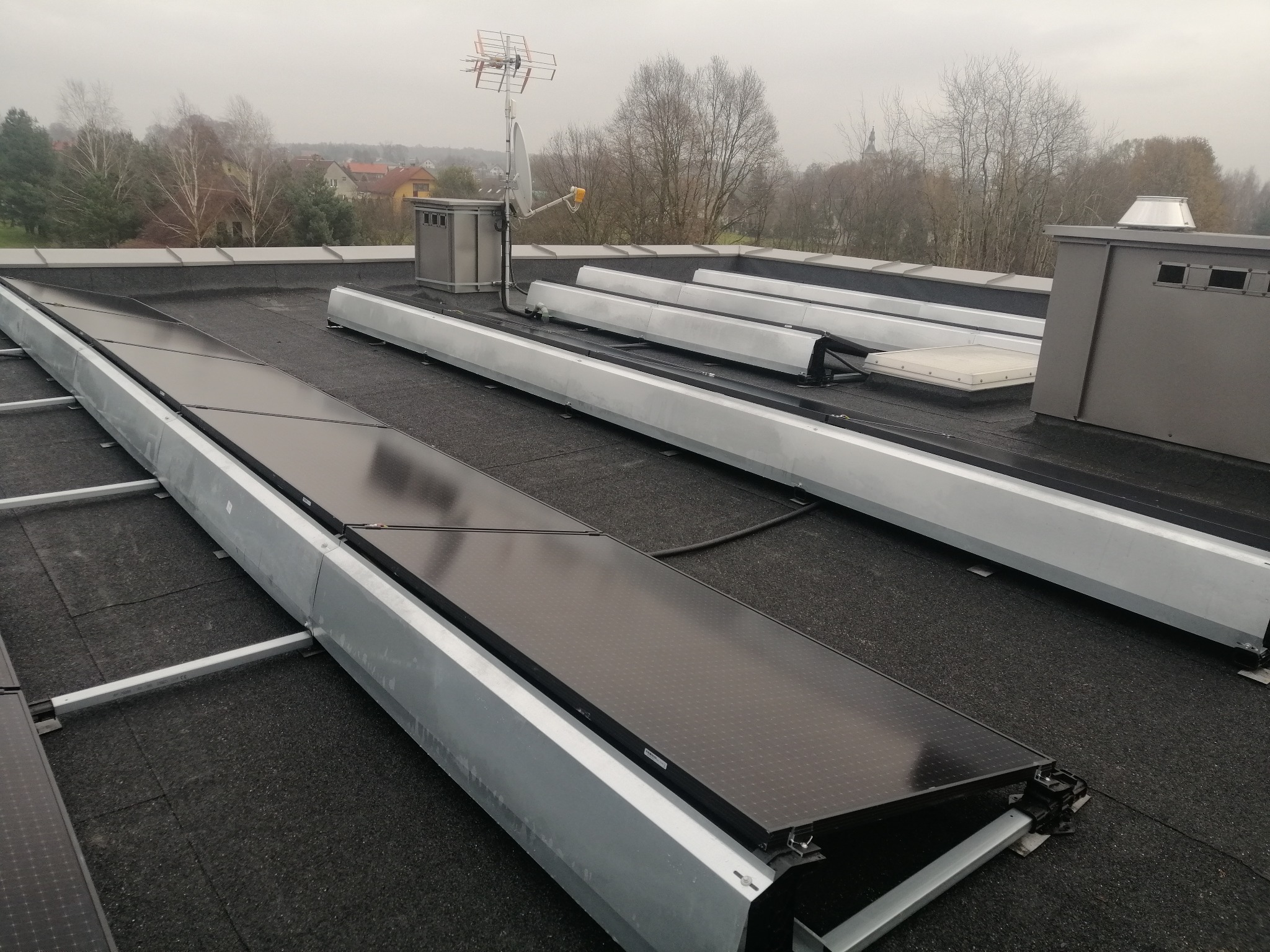 Montaż paneli fotowoltaicznych na płaskim dachu z widoczną konstrukcją wsporczą i elementami wentylacji, w tle zabudowa jednorodzinna i pochmurne niebo.