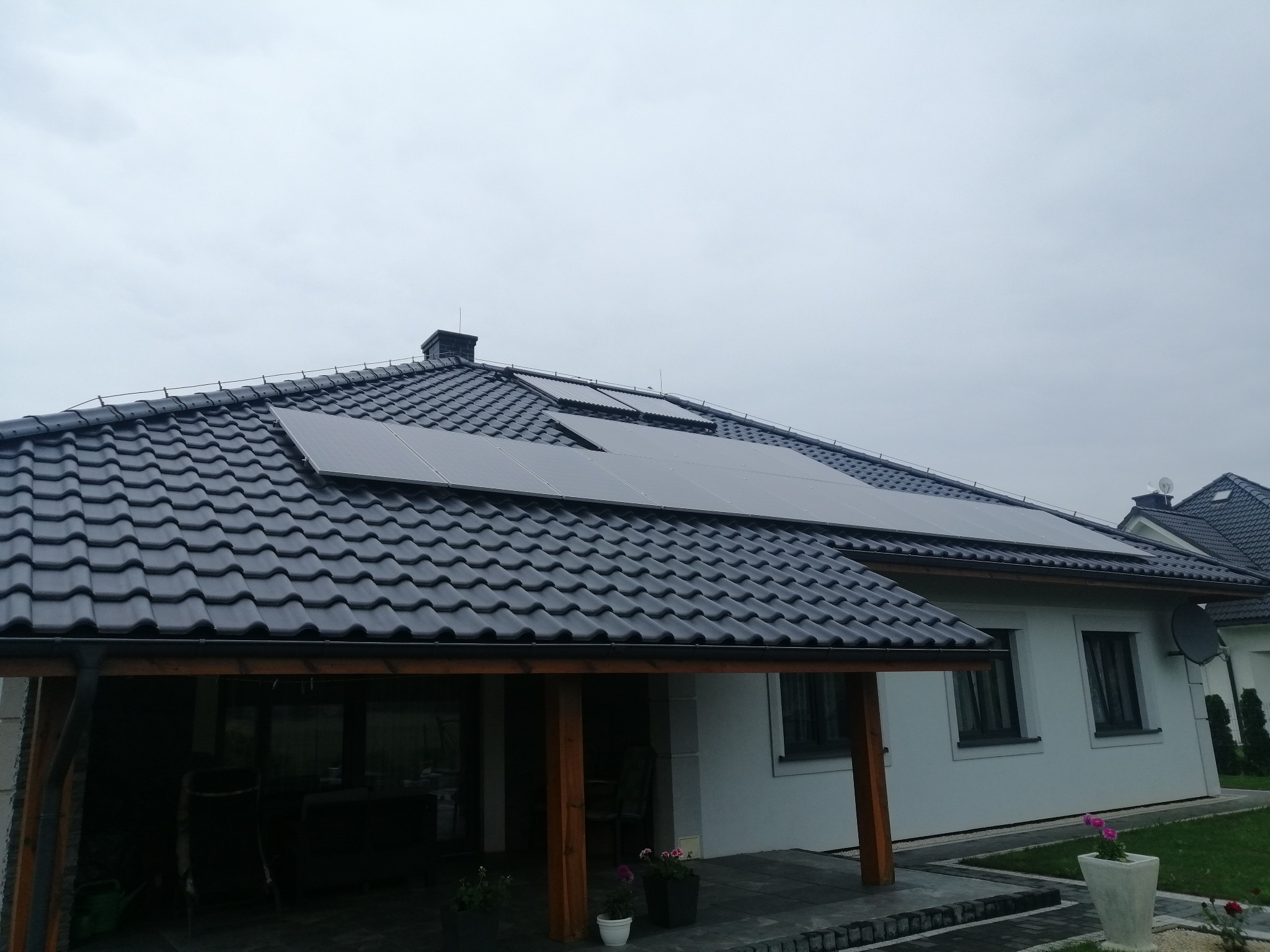 Cztery panele słoneczne na dachu domu z ciemnej dachówki, widok z poziomu tarasu z drewnianymi słupami i kwiatami w donicach, pochmurne niebo w tle.