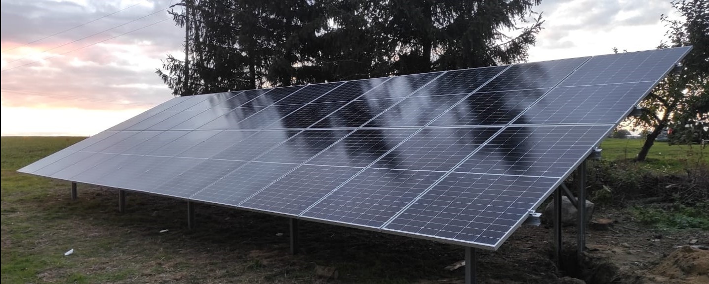 Instalacja fotowoltaiczna na gruncie, widoczne panele na metalowej konstrukcji, w tle drzewa i zachmurzone niebo o zmierzchu.