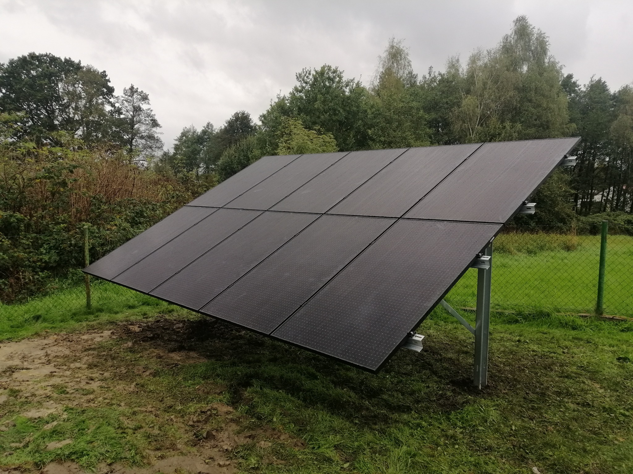 Czarny panel fotowoltaiczny ustawiony na metalowej konstrukcji na zewnątrz, widoczne drzewa w tle i pochmurne niebo.
