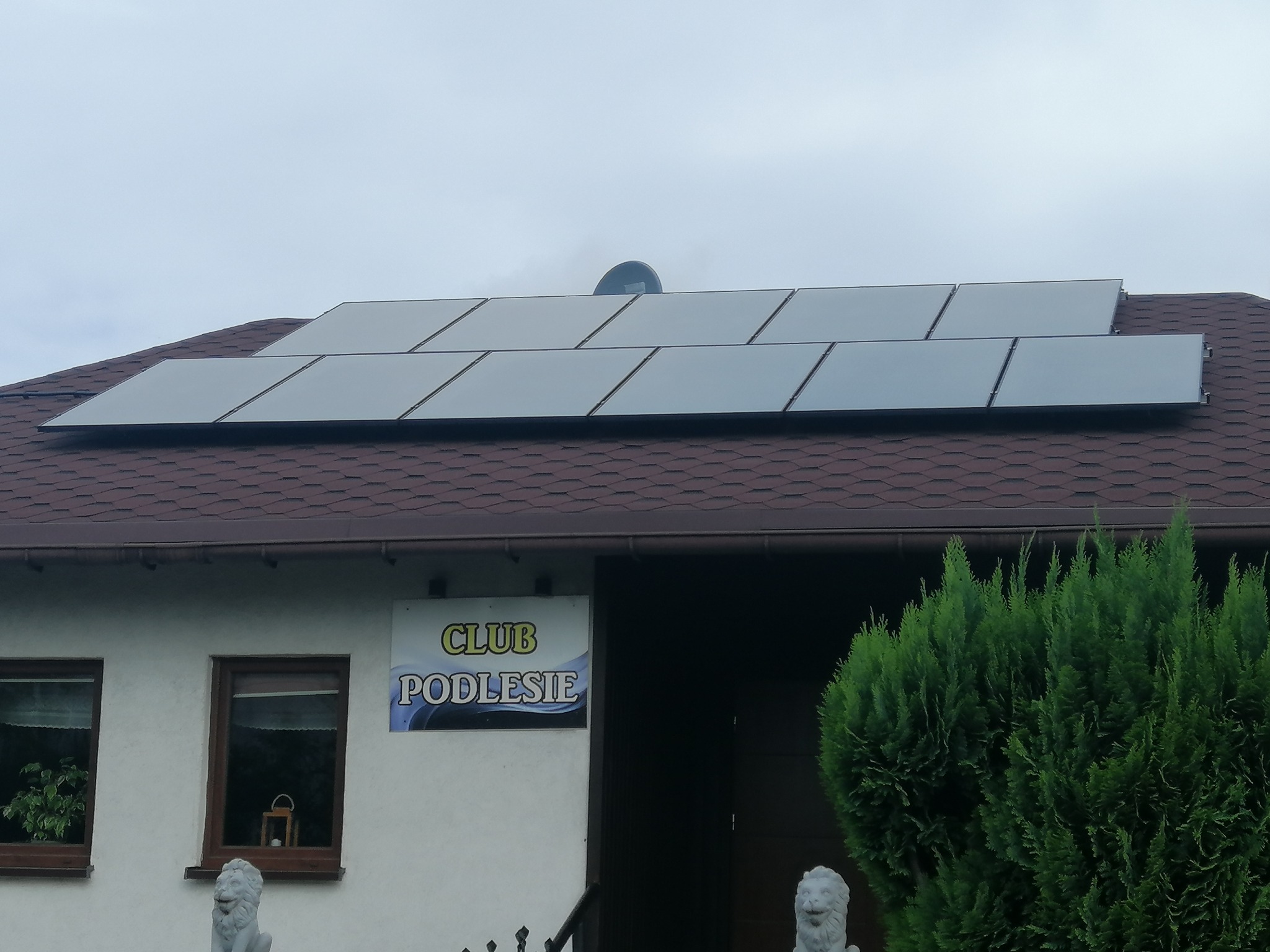 Panele słoneczne na dachu budynku z brązową dachówką i szyldem 'Club Podlesie', widoczne także rzeźby lwów.