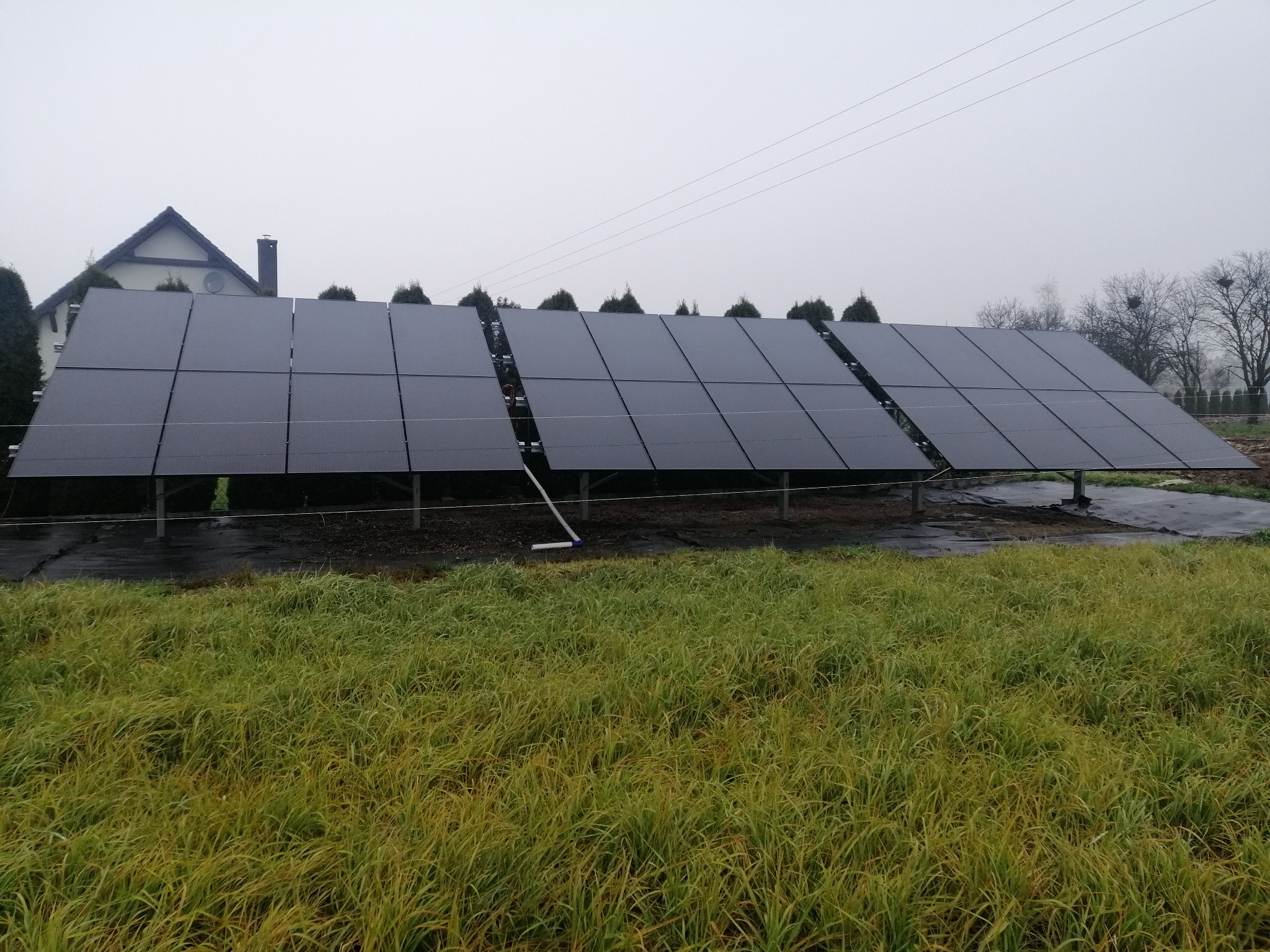 Instalacja paneli słonecznych na trawiastym terenie obok domu w pochmurny dzień. Widoczne trzy rzędy paneli, z tyłu dom z dwuspadowym dachem.