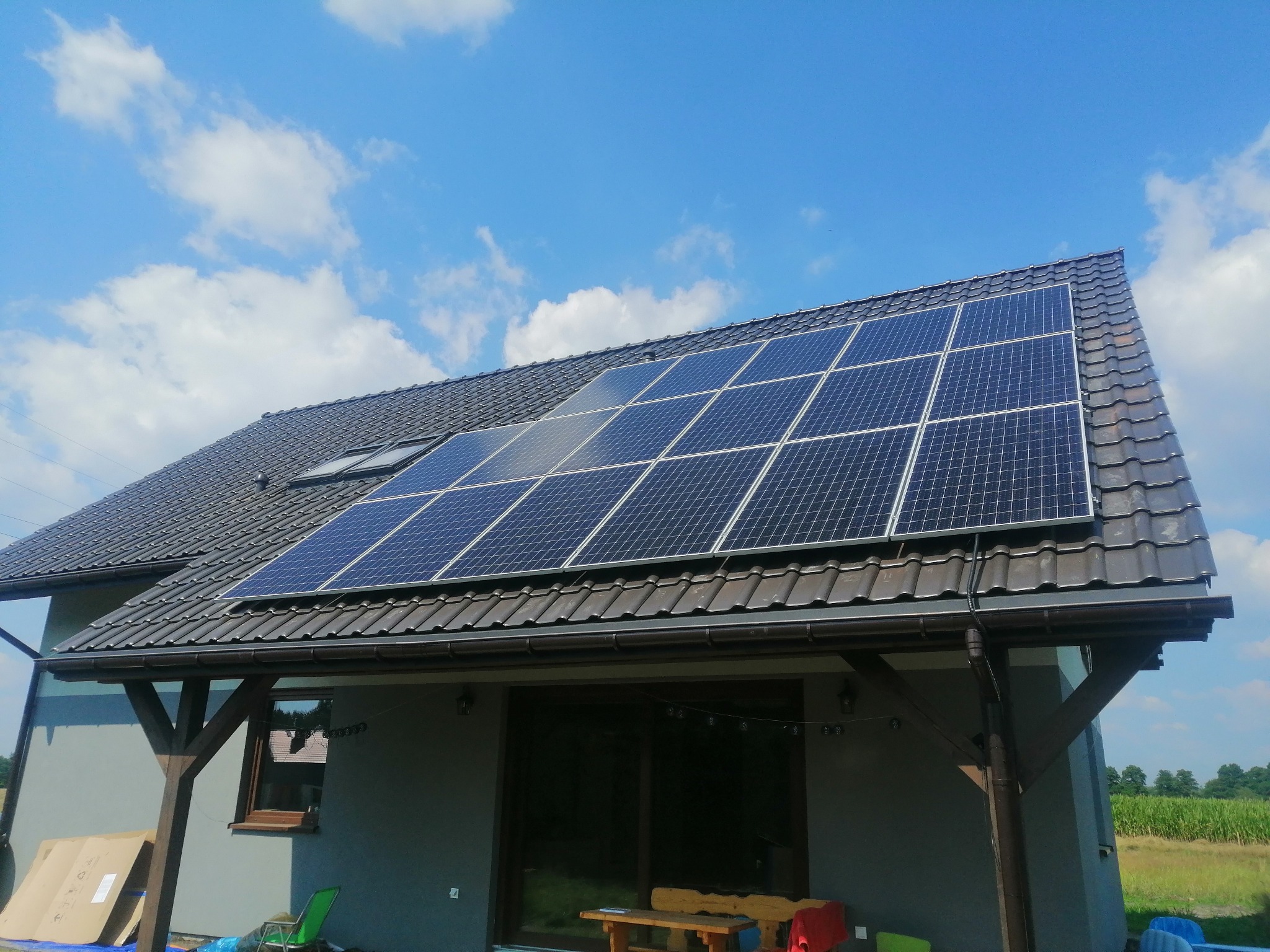 Dom z ciemną dachówką i panelami słonecznymi na dachu, widok z dołu na tle błękitnego nieba z chmurami.