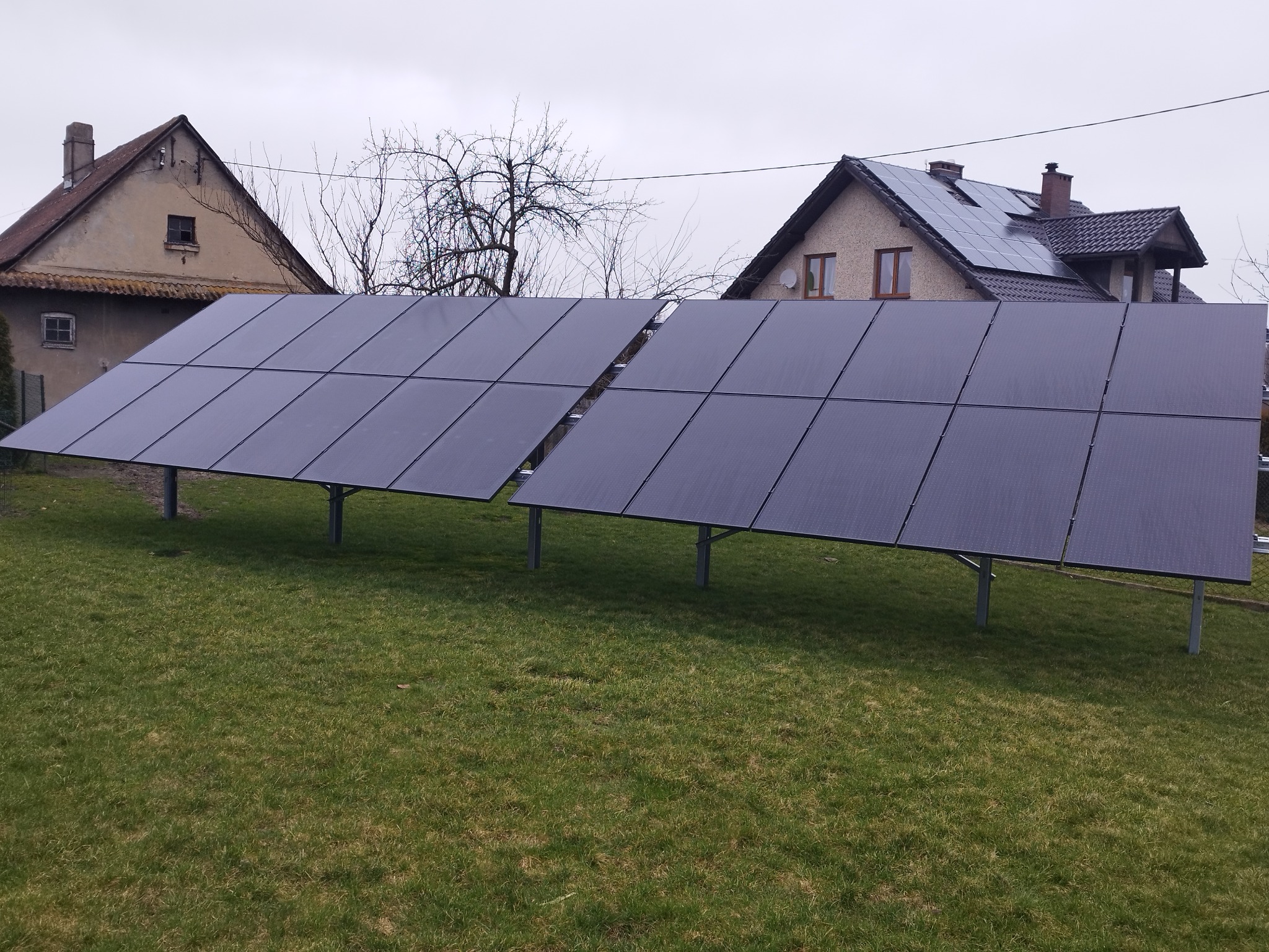 Dwa rzędy paneli słonecznych na trawniku przed domami jednorodzinnymi, część paneli zamontowana również na dachu jednego z budynków.