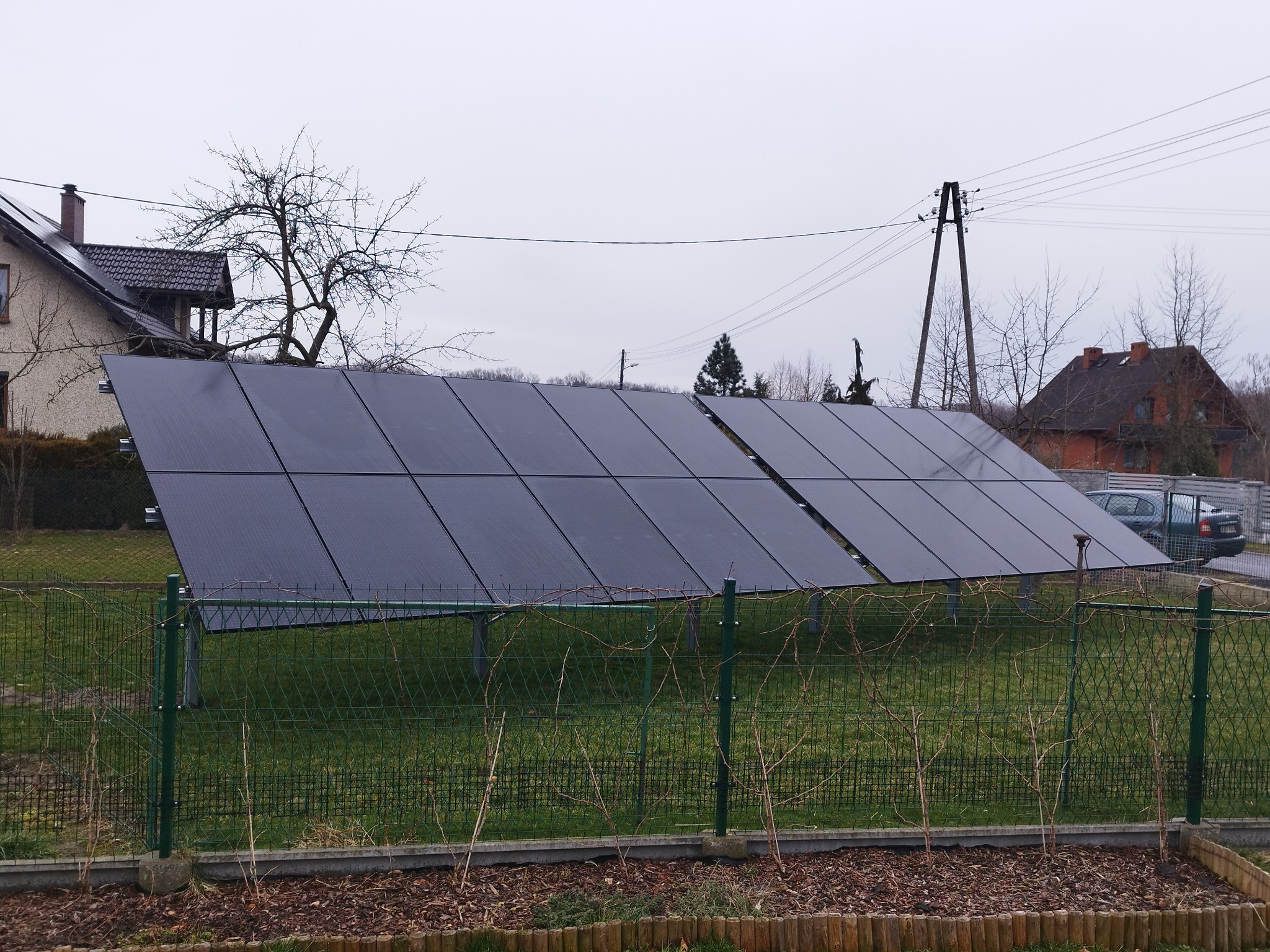 Rząd paneli słonecznych na metalowej konstrukcji, widoczny zza niskiego, zielonego płotu na tle domu i pochmurnego nieba.