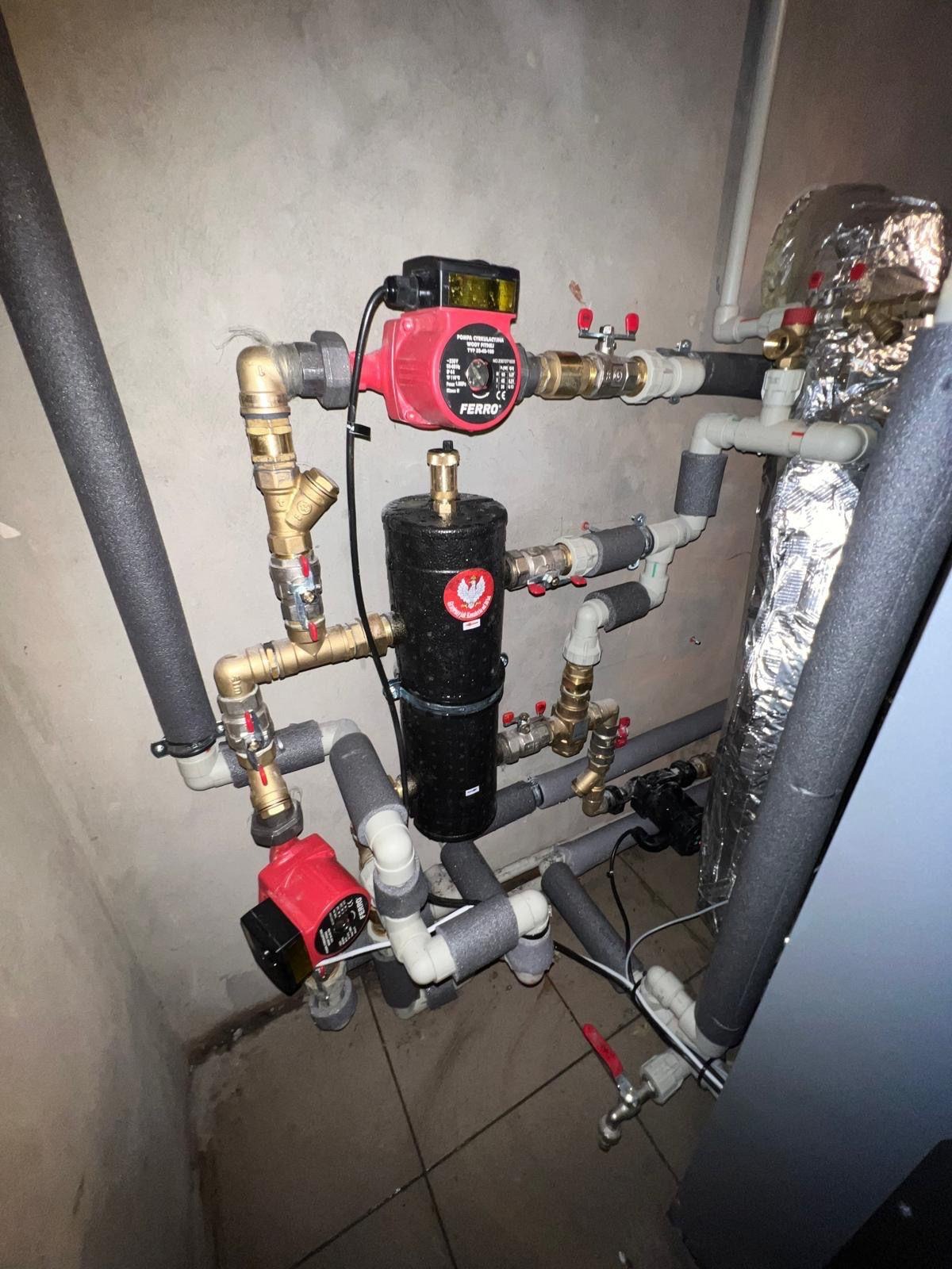 Złożona instalacja hydrauliczna z dwoma czerwonymi pompami obiegowymi marki Ferro, czarnym zbiornikiem z naklejką z orłem w koronie, rurami miedzianymi i izolowanymi rurami PCV, zamontowana...