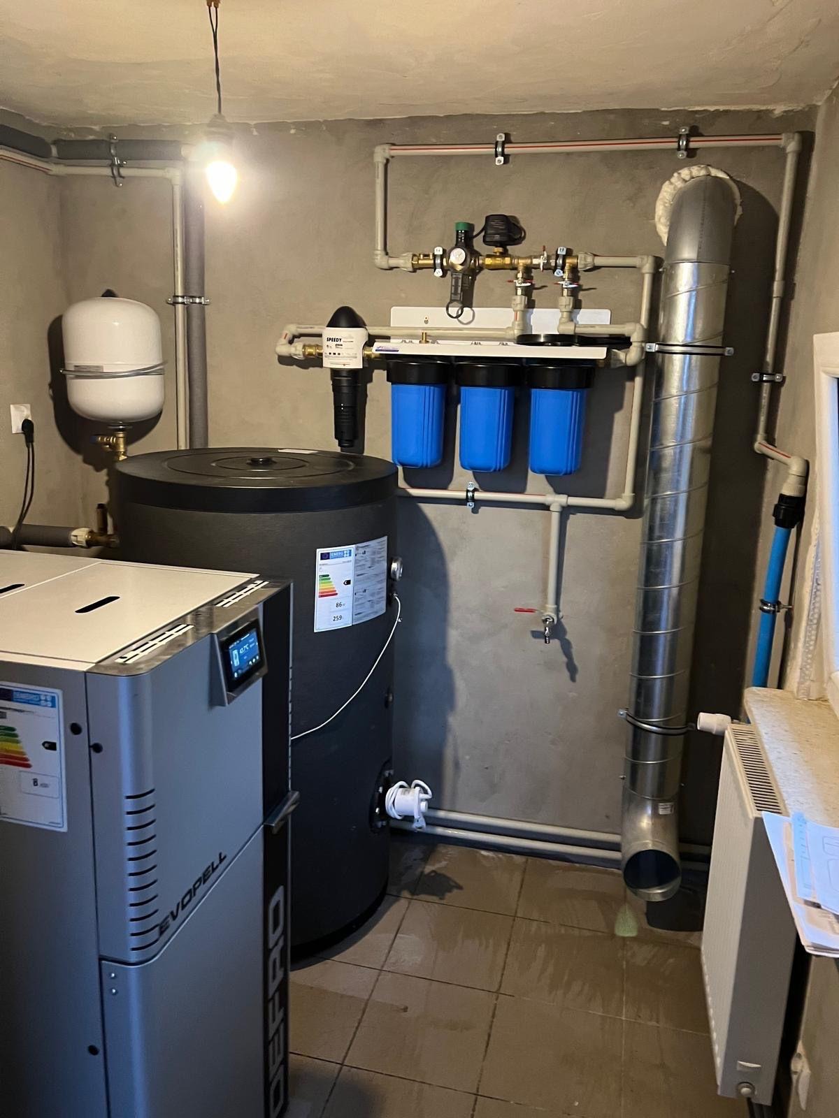 Kotłownia z kotłem na pellet, zbiornikiem buforowym i filtrami wody, widoczne rury instalacji grzewczej, szara ściana i brązowa podłoga z płytek.