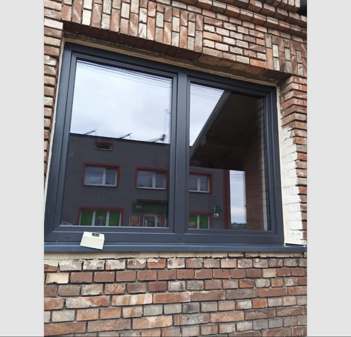 Nowe, grafitowe okno PCV dwuszybowe zamontowane w murze z czerwonej cegły. W szybach odbija się budynek naprzeciwko.