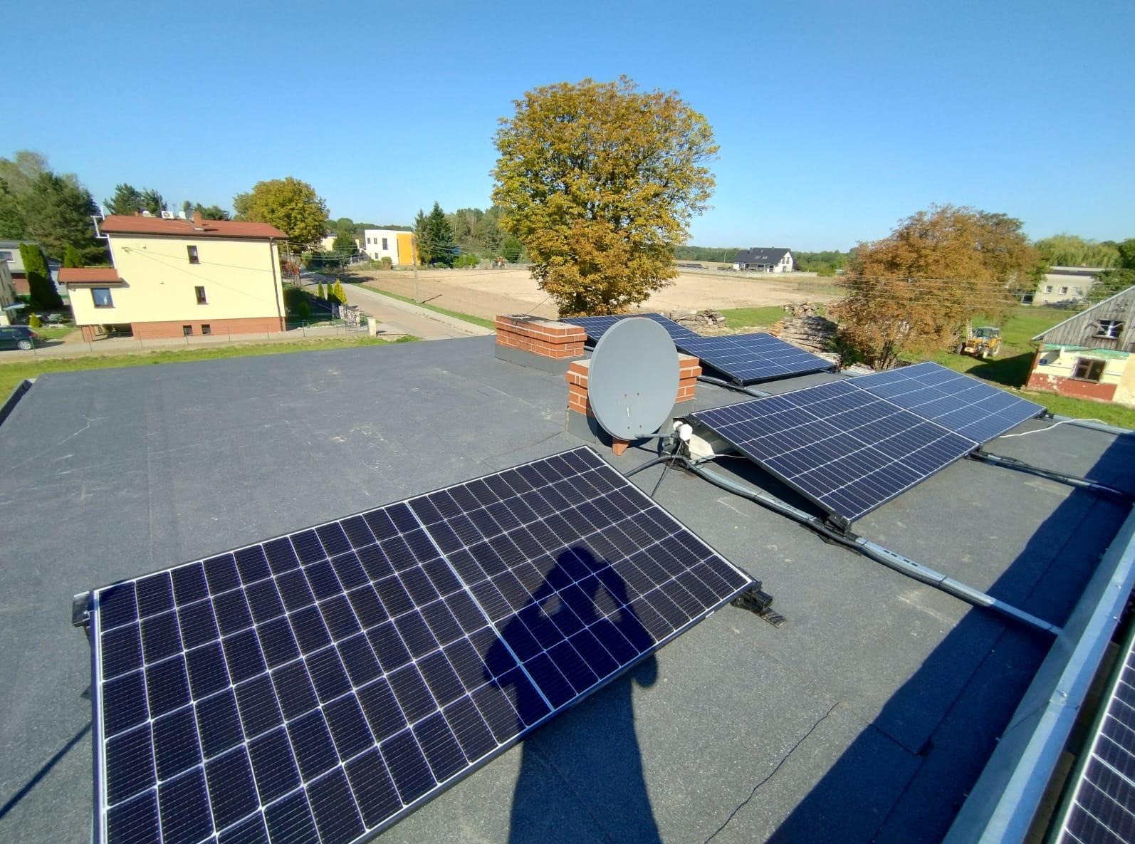 Instalacja fotowoltaiczna na płaskim dachu z widoczną anteną satelitarną i cieniem osoby fotografującej na panelach, w tle zabudowa jednorodzinna i drzewa pod błękitnym niebem.