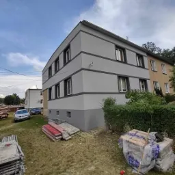 Dwukondygnacyjny budynek po renowacji elewacji w odcieniach szarości z ciemnoszarymi pasami, widoczne rusztowania i materiały budowlane na trawniku.