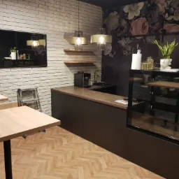 Wnętrze kawiarni z drewnianymi blatami stolików, ladą z ekspozytorem i dekoracyjną ścianą w kwiaty, oświetlone wiszącymi lampami z abażurami.