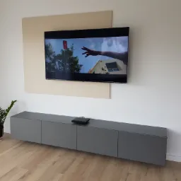 Szara szafka RTV zawieszona pod telewizorem na ścianie z beżowym panelem, minimalistyczny design, podłoga drewniana, roślina w czarnej doniczce.