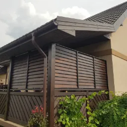 Zadaszony taras z drewnianą zabudową w kolorze brązowym, z widocznymi roślinami i fragmentem elewacji domu.