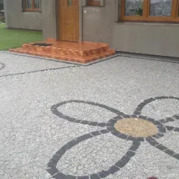 Ułożona nawierzchnia z jasnoszarej kostki granitowej z dekoracyjnymi wzorami: okrąg z koncentrycznymi pierścieniami oraz duży kwiat o żółtym środku i ciemnoszarych płatkach, prowadząca do wejścia...