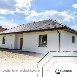 Parterowy dom z ciemnobrązowym dachem, białymi ścianami i oknami z drewnianą ramą, widziany w słoneczny dzień z boku. Na oknie widoczna lampka nocna. Częściowo zagospodarowany teren wokół domu.