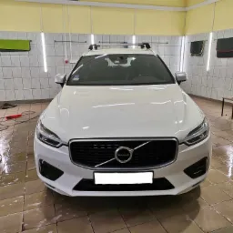 Biały samochód Volvo XC60 po umyciu w myjni, widok z przodu na tle białych płytek i żółtej ściany, z widocznymi śladami wody na podłodze i ręcznikami na ścianie.