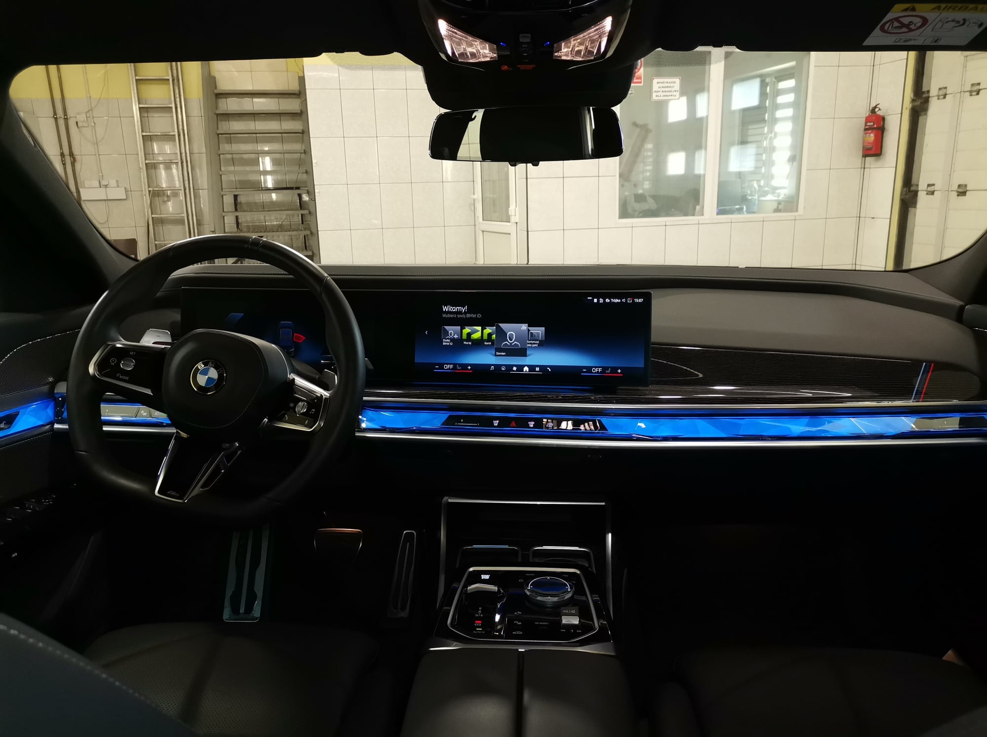 Wnętrze luksusowego samochodu BMW z podświetlaną deską rozdzielczą i ekranem dotykowym, widoczne detale wykończenia i elementy sterowania, tło przedstawia wnętrze myjni samochodowej z drabiną...