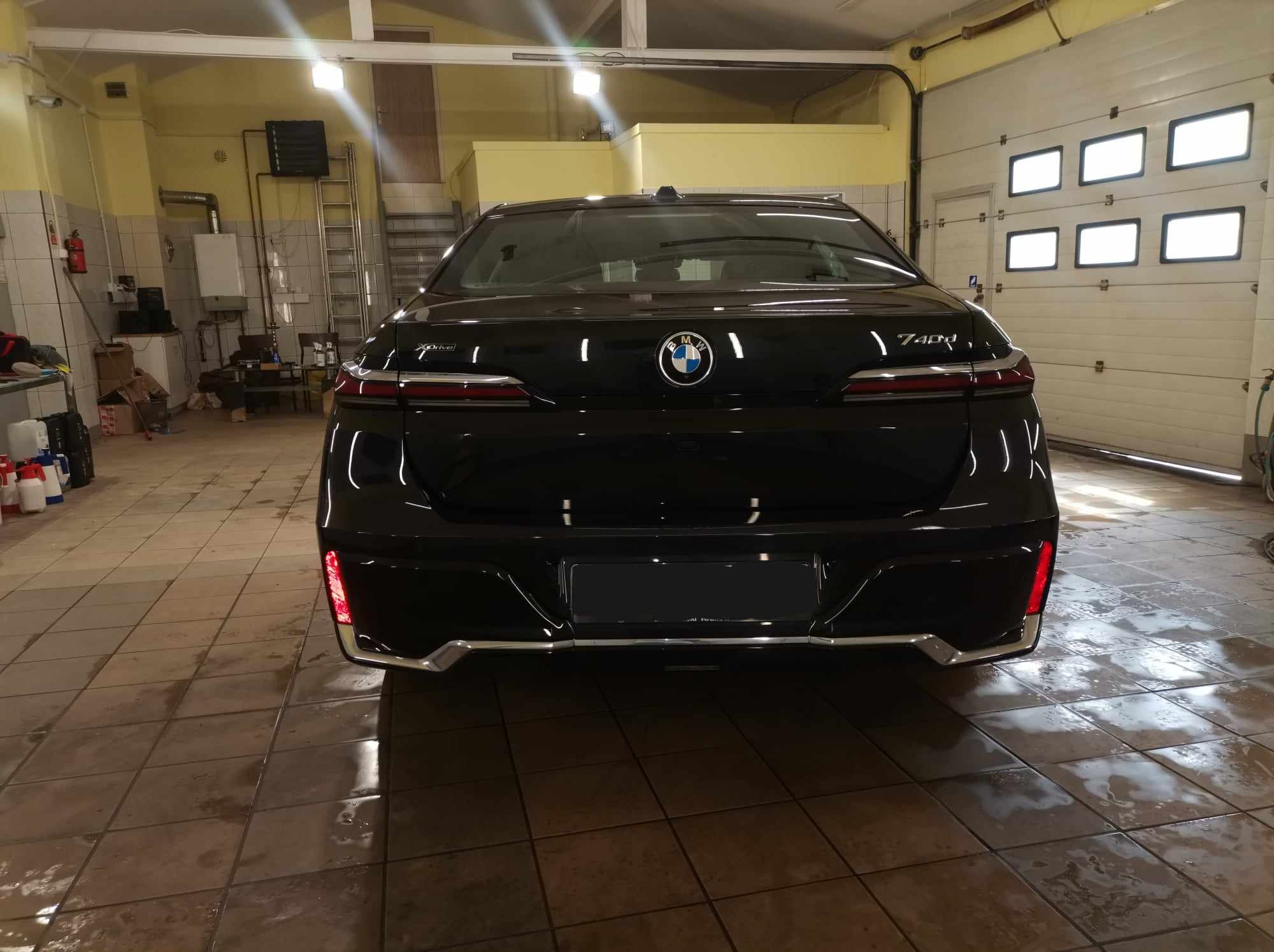 Tył czarnego BMW 740i w garażu z mokrą, brązową podłogą z płytek, widoczny emblemat BMW i światła