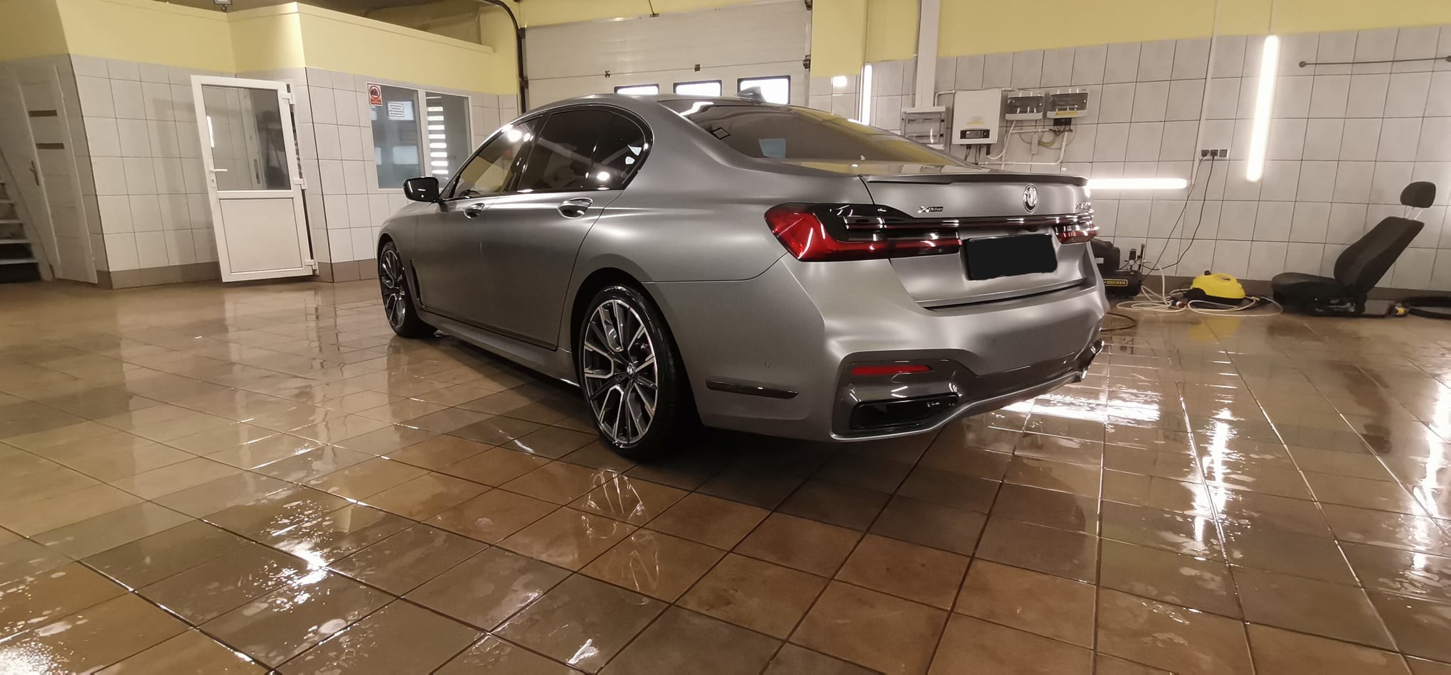 Szary matowy samochód BMW serii 7 po umyciu w myjni, w tle widoczny fotel samochodowy i sprzęt do czyszczenia parowego.