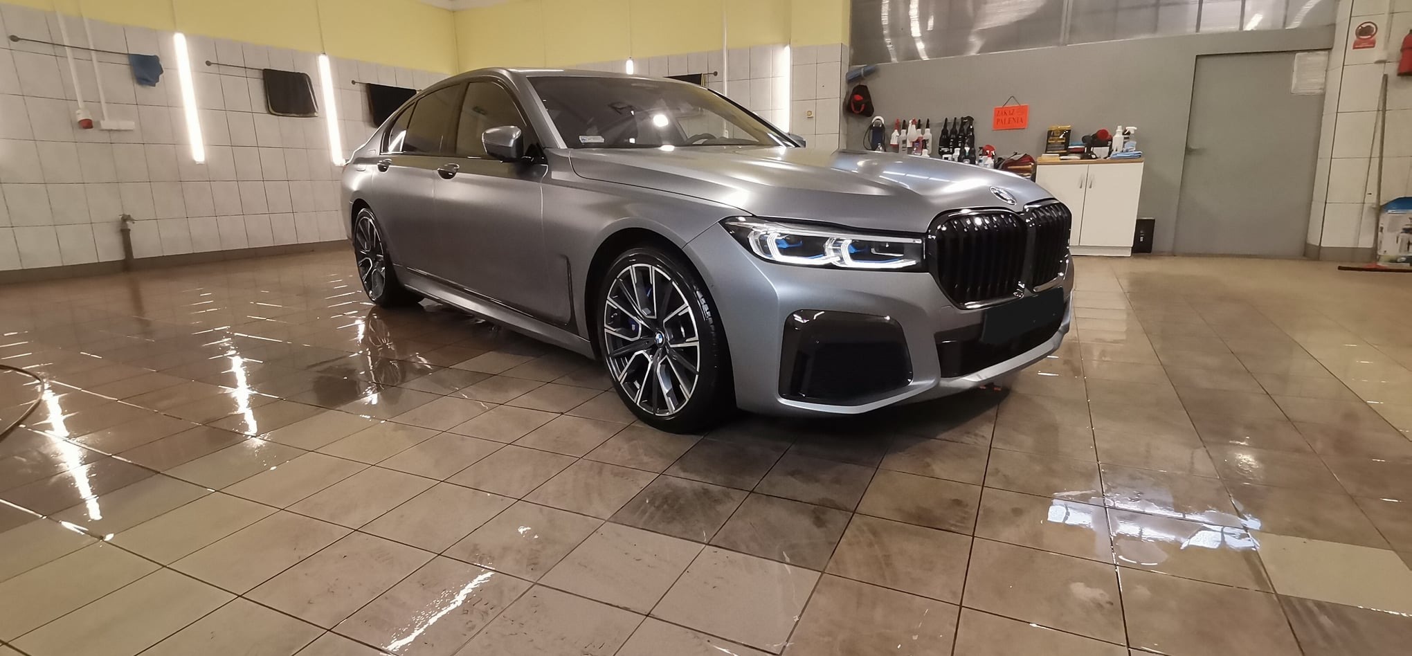 Szary samochód BMW serii 7 po umyciu w myjni, widoczne mokre płytki podłogowe i refleksy światła, w tle regał z kosmetykami samochodowymi.