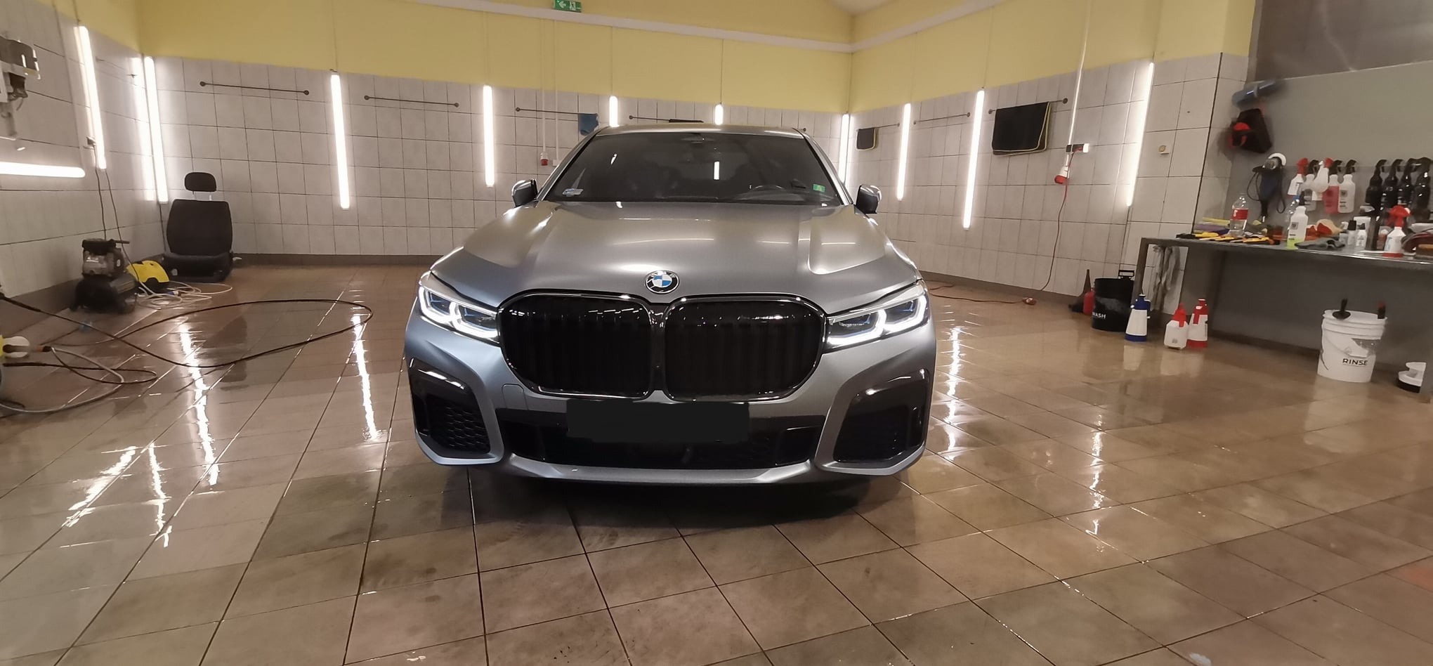 Szary samochód BMW serii 7 po umyciu, widok z przodu na mokrej posadzce myjni, z widocznym wyposażeniem do czyszczenia.