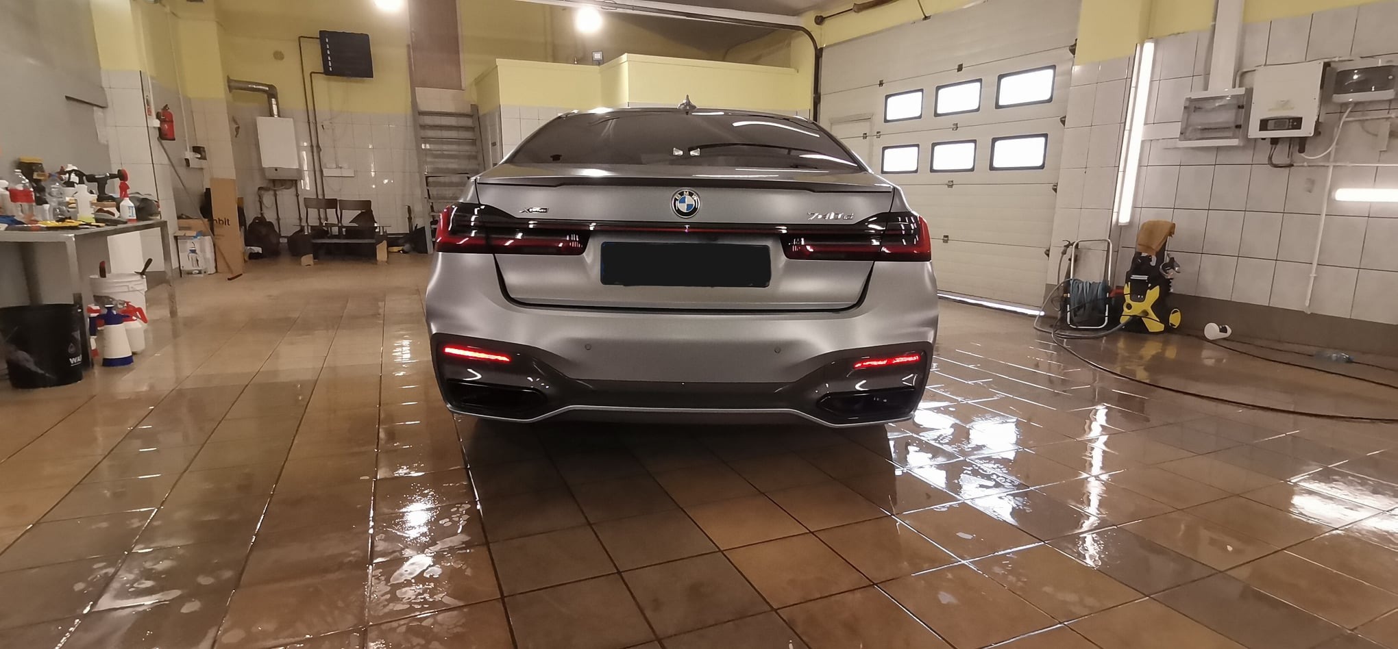 Tył szarego samochodu BMW serii 7 w myjni, widoczny mokry, kafelkowany podłoga i narzędzia do mycia ciśnieniowego.